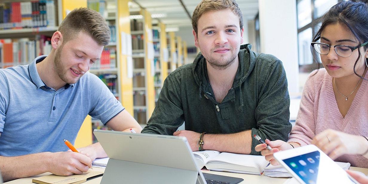 Drei Studierende arbeiten an einem Tisch in einer Bibliothek. Sie benutzen Laptops und Notizbücher, während sie Notizen machen und miteinander kommunizieren. Zwei der Studierenden lachen, was auf eine angenehme und produktive Arbeitsatmosphäre hinweist.